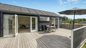 Um deck moderno ao ar livre com área de refeições, um guarda-sol grande e uma porta de vidro deslizante que dá para o interior.