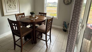 A dining area with a round table and chairs, a ceiling light fixture, a clock, and a window with a view of the outdoors. - Golf, Beach & Great Local Eats! This condo unit is minutes is away from it all. (Breadalbane)