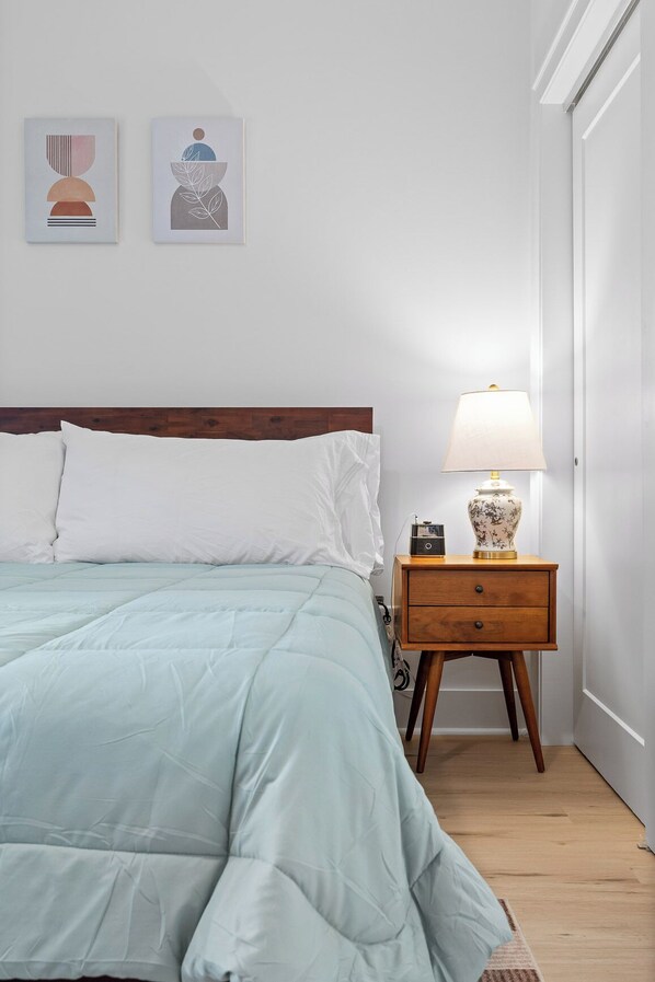 A neatly made bed with a light blue comforter, a wooden nightstand with a lamp, and two framed artworks on the wall.