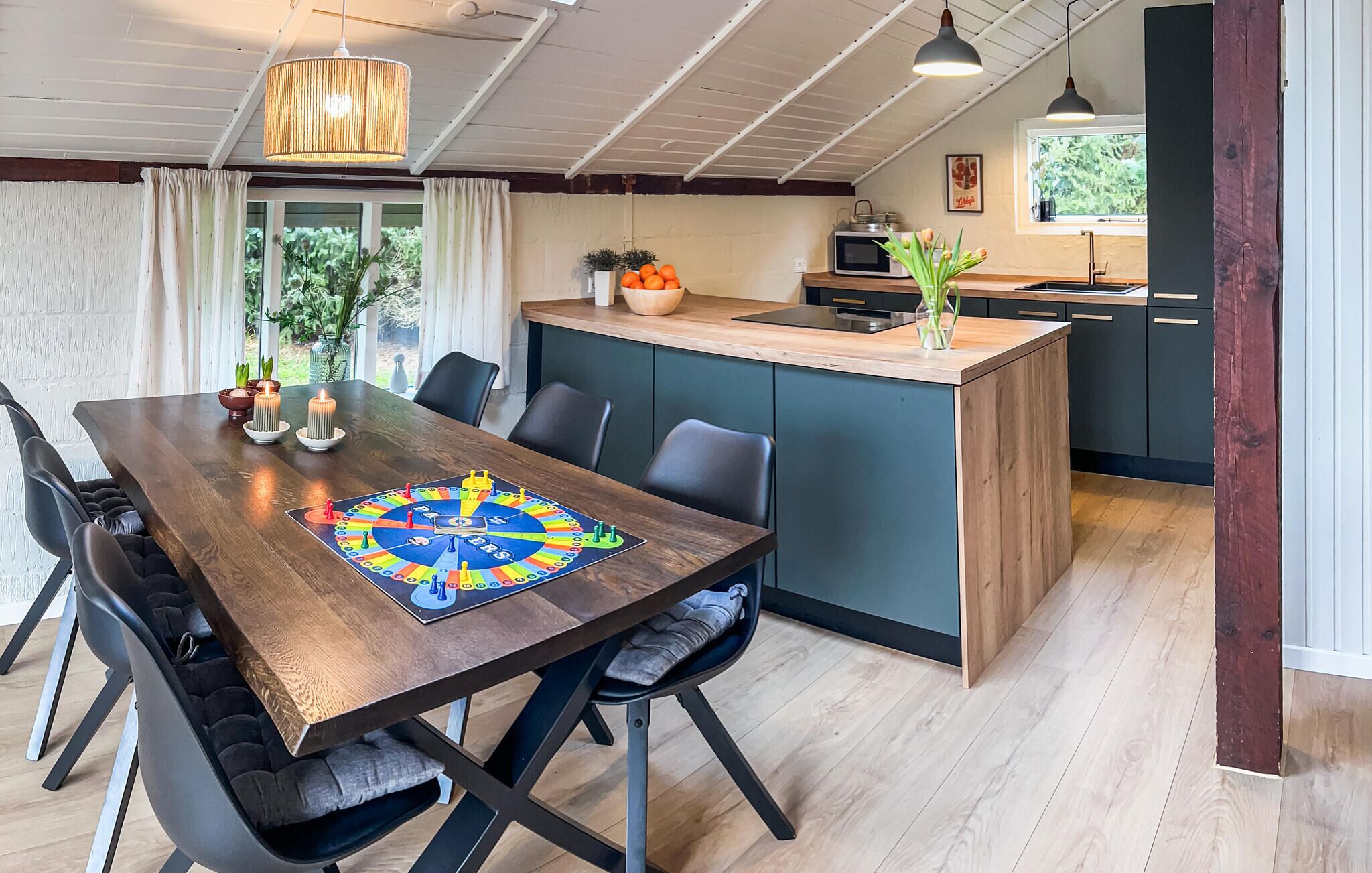A dining area with a wooden table, chairs, and a board game. There is a kitchen area with a microwave and a sink.