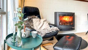 Une pièce chaleureuse avec un fauteuil en cuir noir, une petite table ronde ornée d’un vase et de tasses, et un pouf noir. On aperçoit un poêle à bois au fond de la pièce.