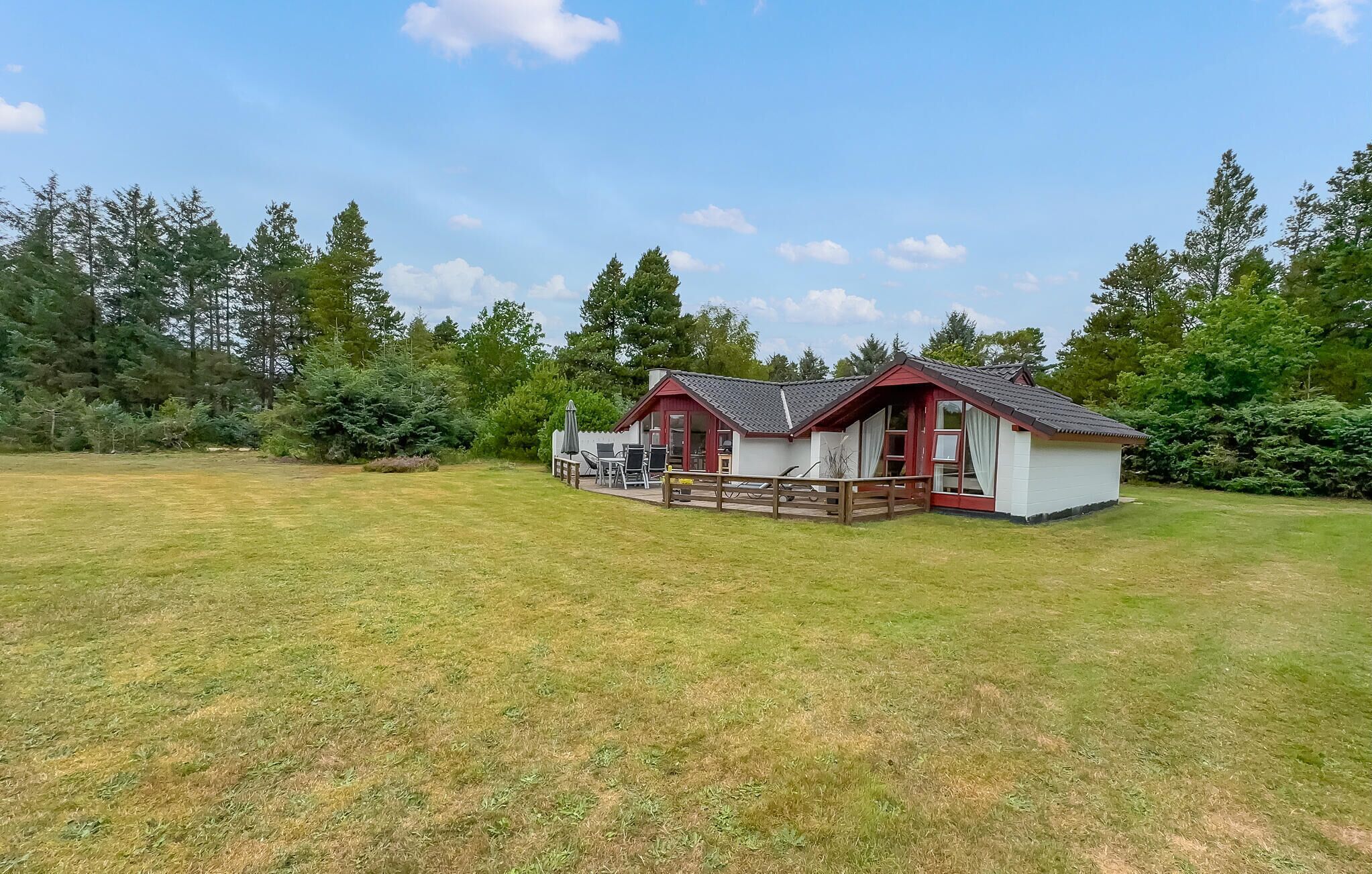 A small, single-story cabin with a red roof and white walls, surrounded by a grassy field and dense trees.
