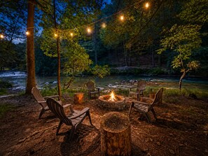 An outdoor seating area with chairs and a fire pit, surrounded by trees and a river.
