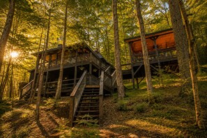 A wooden cabin with a staircase leading to a deck, surrounded by trees and natural light.