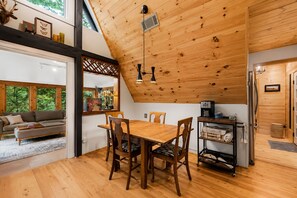 A cozy room with a wooden ceiling, a dining table with chairs, a sofa, and a coffee machine.