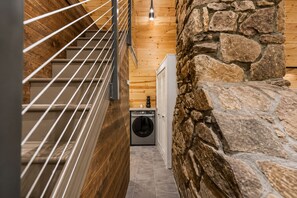 A narrow hallway with a wooden staircase, a stone wall, and a washing machine.