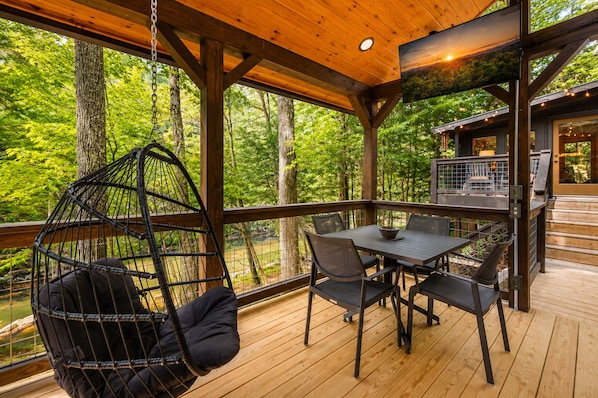 A wooden deck with a hanging chair, a table with chairs, and a view of the forest.