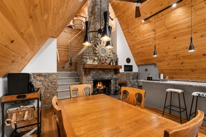 Une cuisine chaleureuse avec une cheminée en pierre, un plafond en bois et un coin repas comprenant une grande table et des chaises en bois.