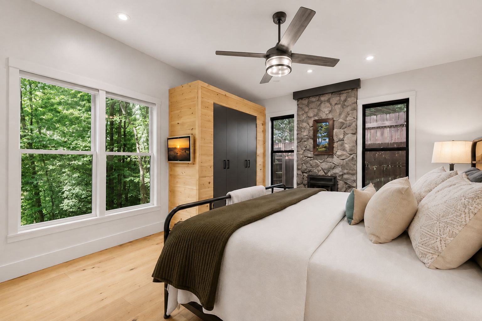 A bedroom with a bed, a ceiling fan, a stone fireplace, and large windows.