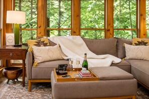 Un salon chaleureux avec un canapé d’angle, une table basse sur laquelle sont posés des livres et une bouteille, une lampe, et une vue sur une forêt à travers de grandes fenêtres.