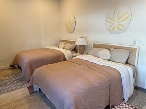 A hotel room with two beds, each with a brown bedspread, white linens, and decorative pillows. There is a nightstand with a lamp and a wall-mounted artwork.