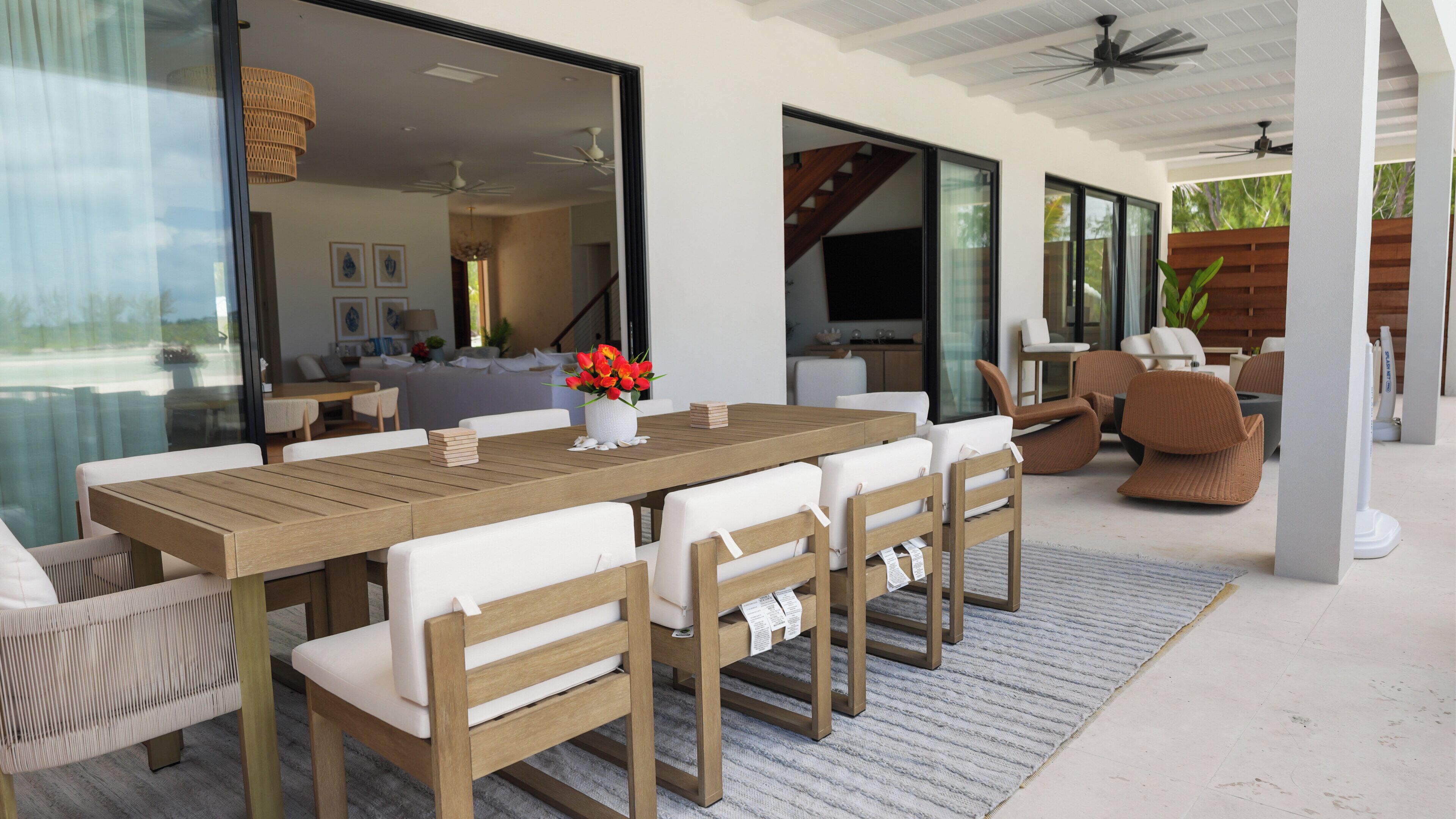 Un amplio espacio de comedor al aire libre con una mesa de madera y sillas blancas acolchadas, un pequeño jarrón blanco con flores rojas y vistas al interior a través de puertas de cristal.