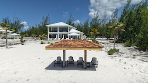 Une propriété située en bord de mer, comprenant un bâtiment à deux étages, un espace extérieur abrité avec des chaises, et une plage de sable.