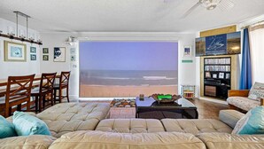 A living room with a large window overlooking the beach, a ceiling fan, a bookshelf, and a dining area with wooden chairs.