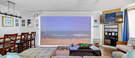 A living room with a large window overlooking the beach, a ceiling fan, a bookshelf, and a dining area with wooden chairs.
