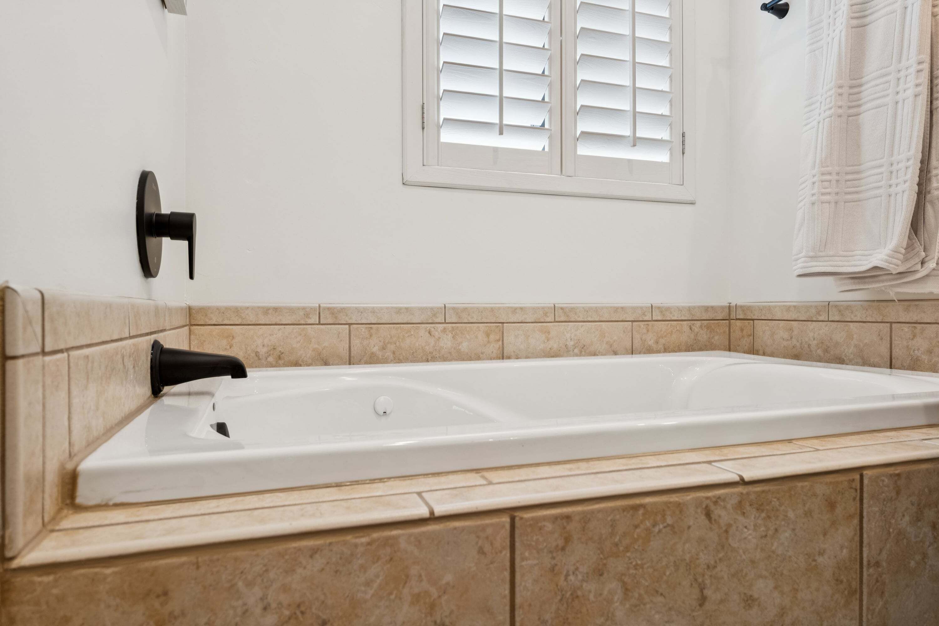 A bathroom with a white bathtub, beige tiled walls, a window with white shutters, and a towel hanging on the wall.