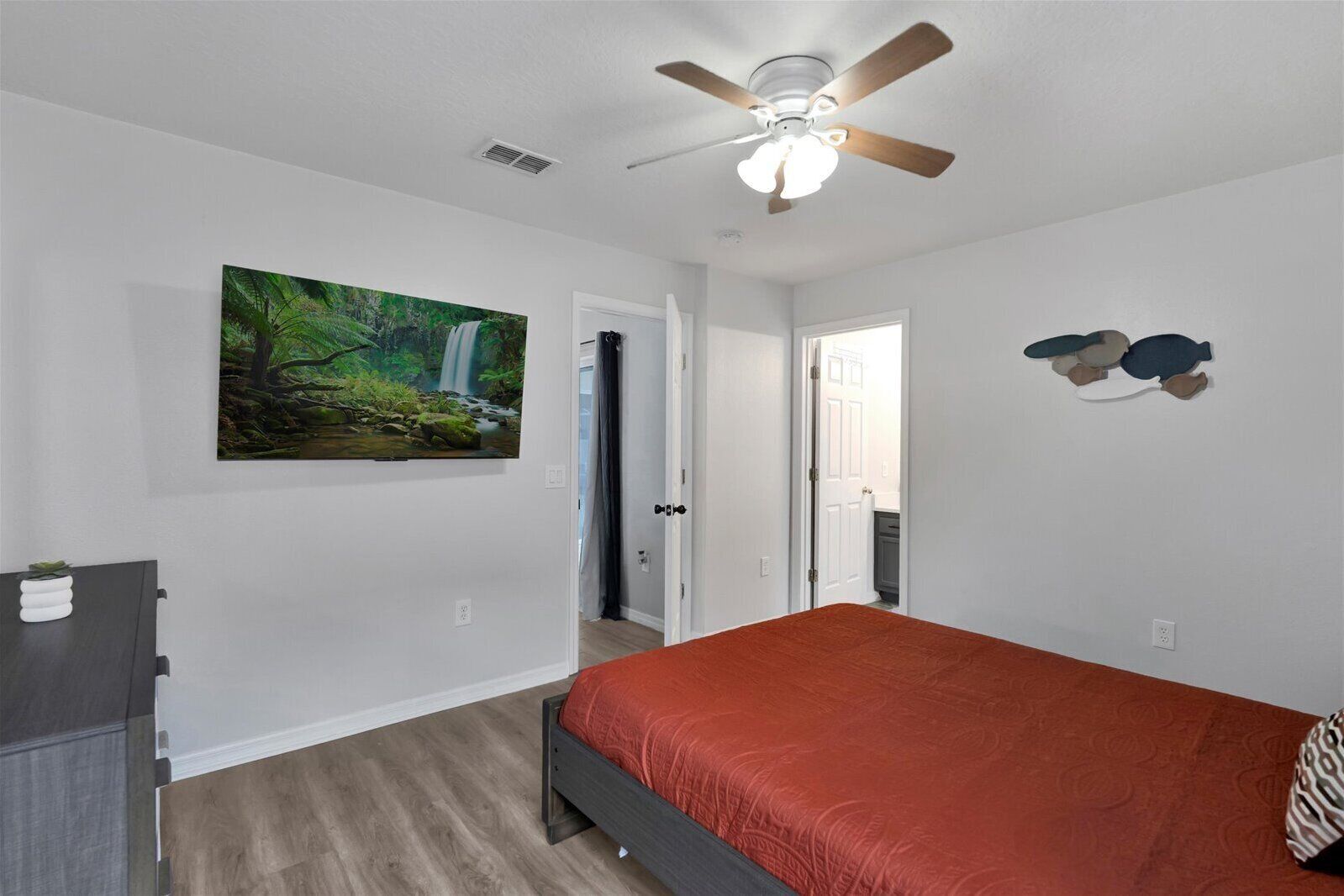 A bedroom with a bed, a ceiling fan, a painting of a waterfall, and a dresser.