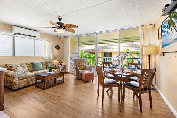 Un salón espacioso con ventilador de techo, un comedor con muebles de madera y ventanas grandes que ofrecen vistas al exterior.