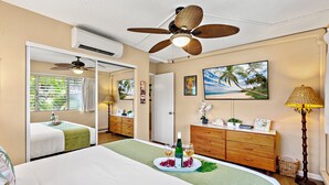 A hotel room with a bed, a dresser, a mirror, a ceiling fan, a painting of a beach, and a tray with wine glasses and fruit. - Tropical stay with full kitchen (Kihei)