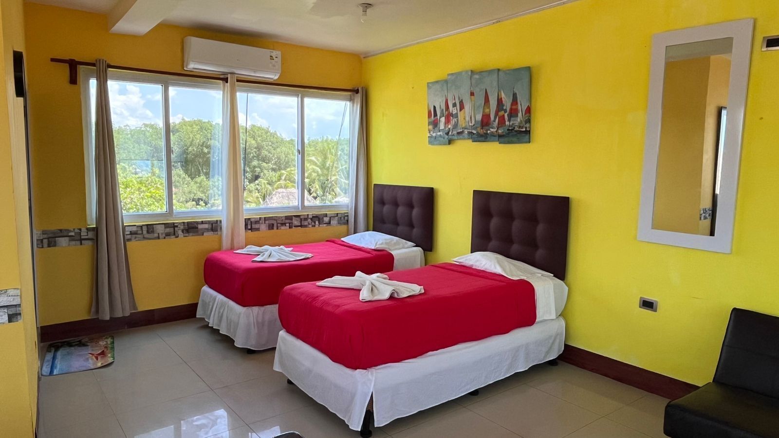 A hotel room with two beds, a large window with curtains, a painting on the wall, and a mirror.