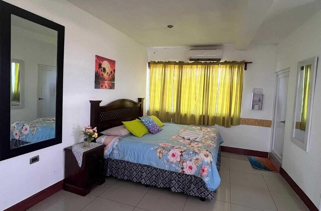 Un dormitorio con cama, mesita de noche, espejo, ventana con cortinas y un equipo de aire acondicionado.