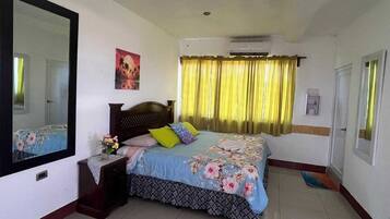 Un dormitorio con cama, mesita de noche, espejo, ventana con cortinas y un equipo de aire acondicionado.