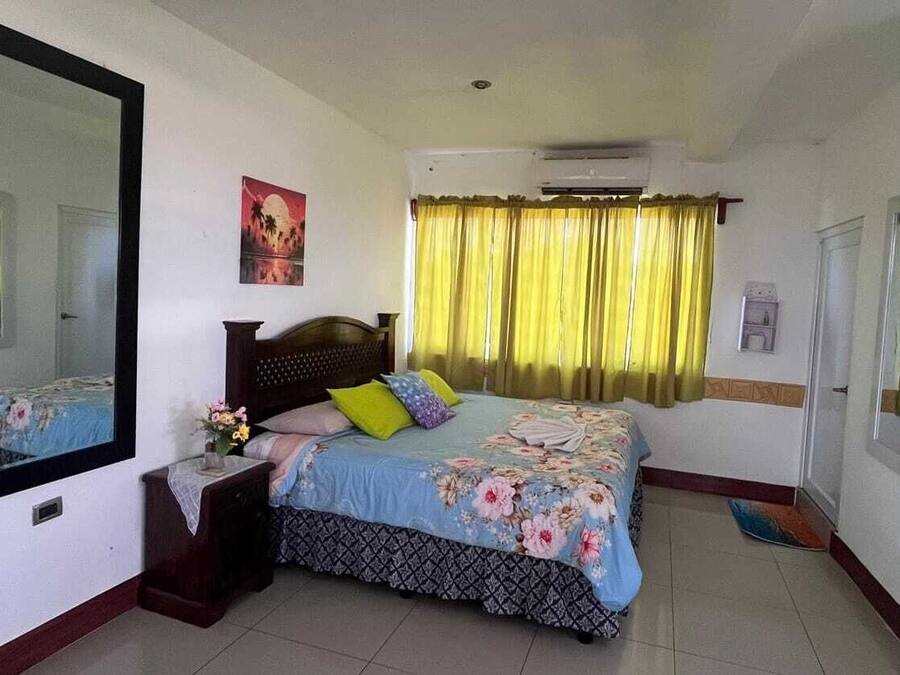 Un dormitorio con cama, mesita de noche, espejo, ventana con cortinas y un equipo de aire acondicionado.