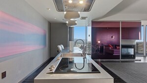 A modern hotel room with a large window, a fireplace, a dining area, and a view of the cityscape.