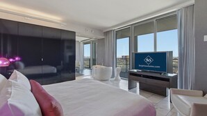 A modern hotel room with a large bed, a flat-screen TV, a bathtub, and a view of the city.