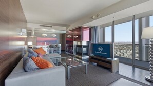 A modern hotel room with a large TV, a sofa, a glass coffee table, and a view of the city.