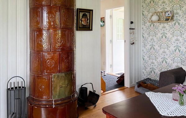 Una habitación con un pilar de madera, un cuadro enmarcado en la pared, un pequeño estante con objetos decorativos, una mesa de madera con mantel de encaje, y una cama para perro en la esquina.