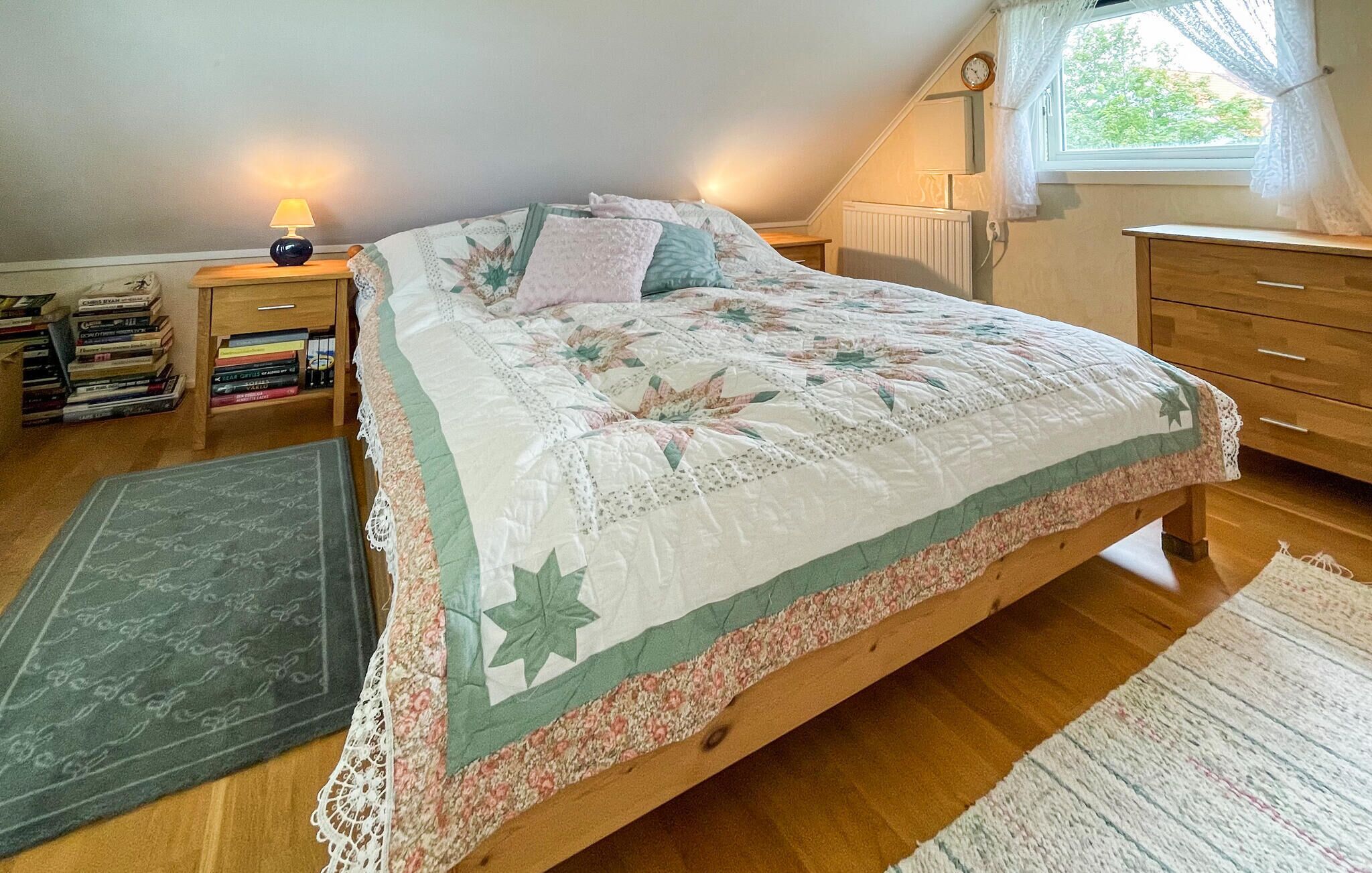 A bedroom with a bed, a bedside table with a lamp, a dresser, a window with curtains, and a radiator.