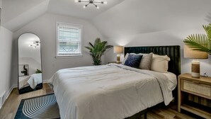 A bedroom with a large bed, a ceiling light fixture, a window with blinds, a bedside table with a lamp, and a mirror. - Modern Home 3 Minutes from Airport! (Cheektowaga)