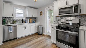 A modern kitchen with white cabinets, a stainless steel dishwasher, a trash can, a microwave, and a stove. - Modern Home 3 Minutes from Airport! (Cheektowaga)