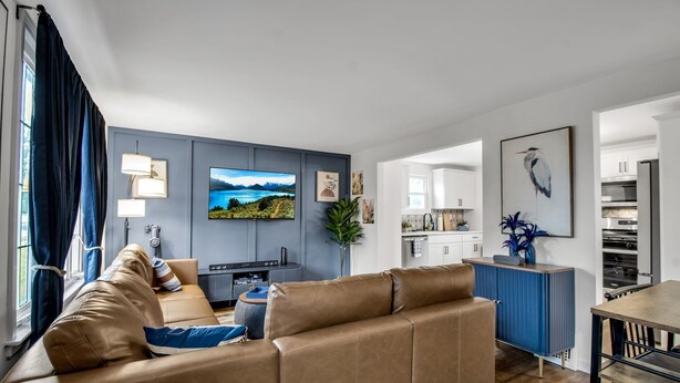 A modern living room with a large brown leather sofa, a flat-screen TV mounted on the wall, and a dining area with a wooden table and chairs. - Modern Home 3 Minutes from Airport! (Cheektowaga)