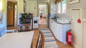 Eine schmale Küche mit Esstisch und Stühlen, einem Herd, einem Backofen und einem Kühlschrank. An der Wand hängt ein Feuerlöscher und es gibt ein Fenster mit Vorhängen.