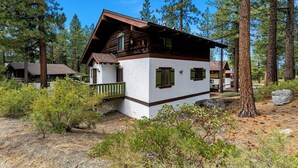 Una cabaña de paredes blancas y detalles marrones, rodeada de pinos y terreno seco.