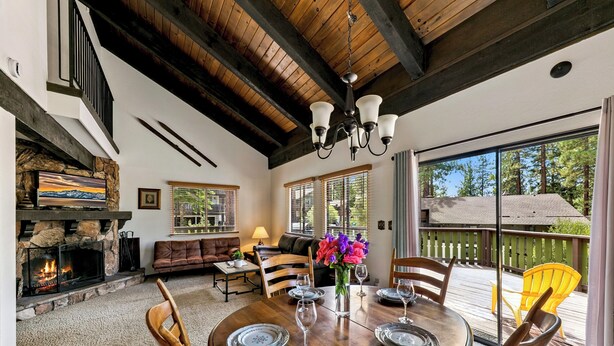 Un comedor acogedor con una mesa de madera dispuesta para cuatro personas, rodeada de sillas de madera. En el fondo, se aprecia una chimenea con revestimiento de piedra, y una ventana grande ofrece vistas al exterior.