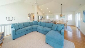 A spacious living room with a large blue sectional sofa, a dining area with a wooden table and chairs, and a kitchen in the background.