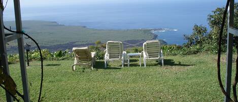 A grassy area with three white lounge chairs and a small table, overlooking a coastal landscape.