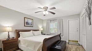 Un dormitorio con cama, mesitas de noche, un banco, ventilador de techo, ventana con cortinas y una puerta.