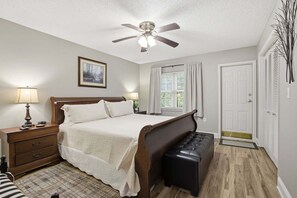 Un dormitorio con cama, mesitas de noche, un banco, ventilador de techo, ventana con cortinas y una puerta.