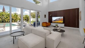 Una sala de estar moderna con un televisor grande, sofás blancos, una mesita y vistas a una piscina y palmeras.