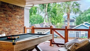 Een biljarttafel op een houten terras met een bakstenen muur op de achtergrond, met uitzicht op een woongebied met huizen en palmbomen.