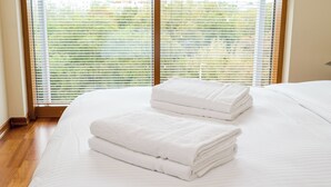 Una cama bien hecha con toallas blancas dobladas y vistas a un paisaje verde a través de amplios ventanales.