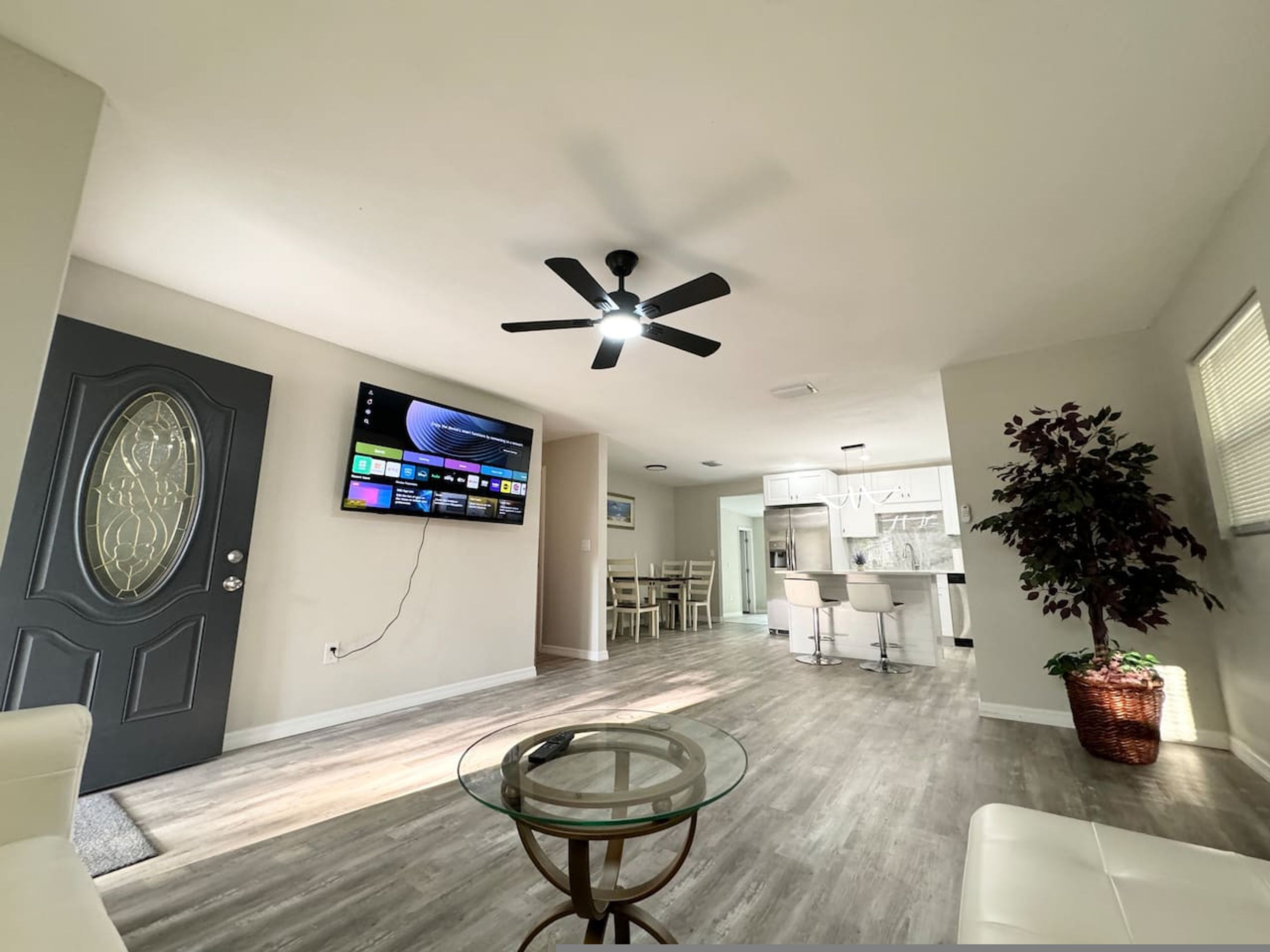 A modern living room with a glass coffee table, a ceiling fan, a flat-screen TV mounted on the wall, and a dining area in the background.