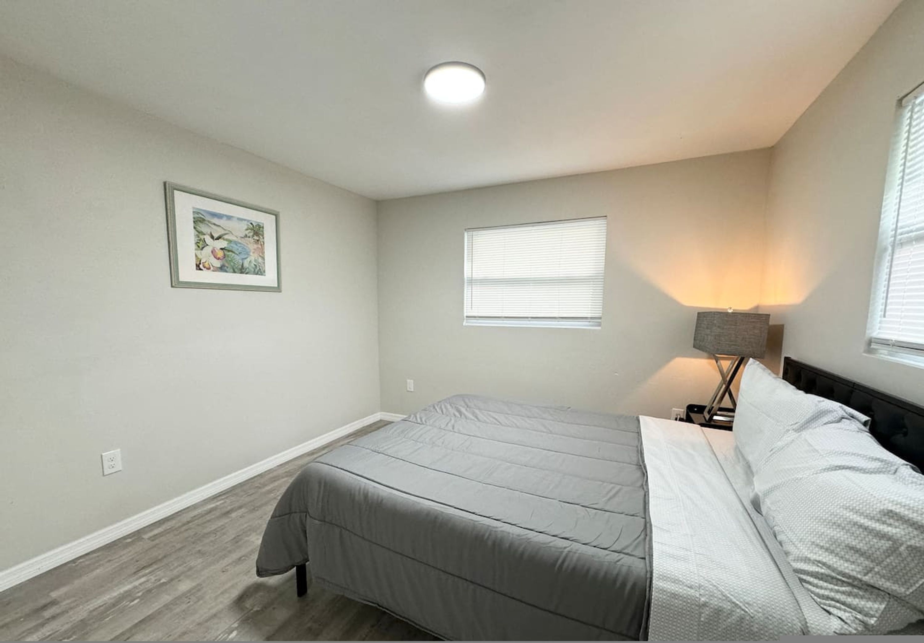 A bedroom with a bed, a nightstand, a lamp, a window, and a wall-mounted picture.