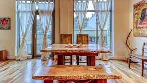 Un espace repas avec une table et des bancs en bois, de grandes fenêtres avec des rideaux et des œuvres d’art accrochées aux murs.