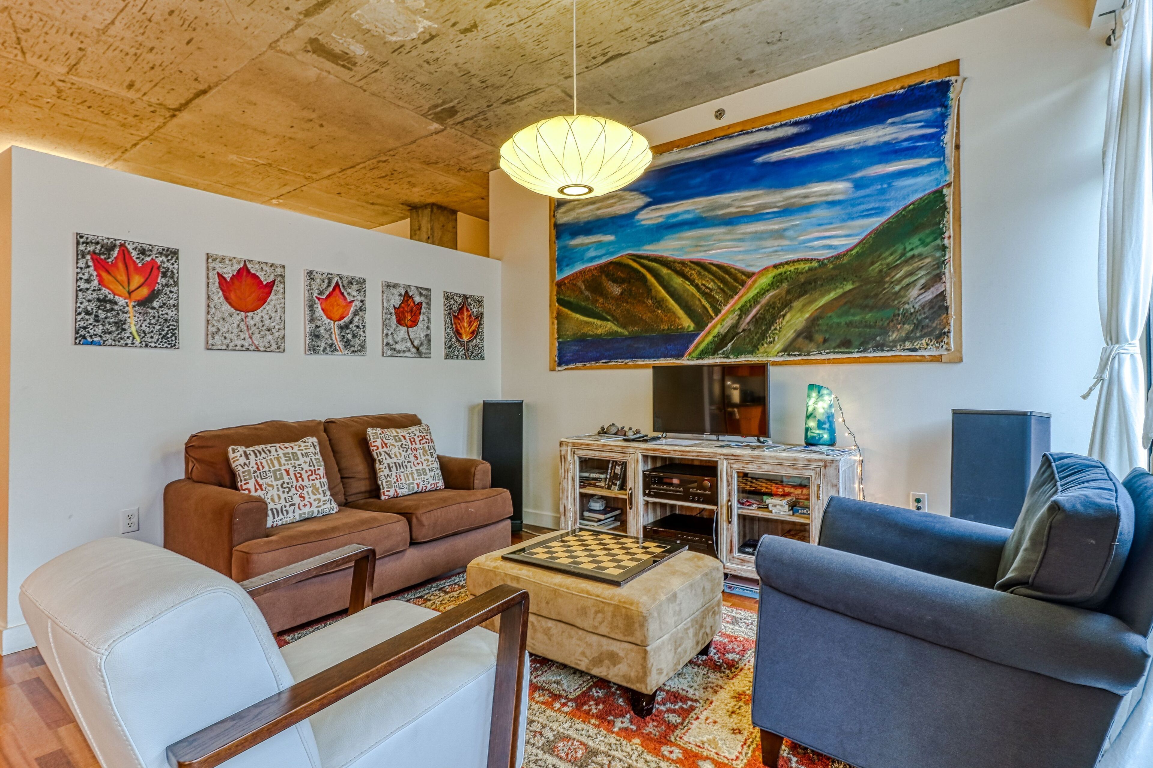Sala de estar com um sofá marrom, uma poltrona branca, uma poltrona cinza, uma mesa de centro e um quadro de uma paisagem.