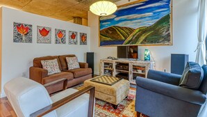 Un salon avec un canapé marron, un fauteuil blanc, un fauteuil gris, une table basse et un tableau représentant un paysage.